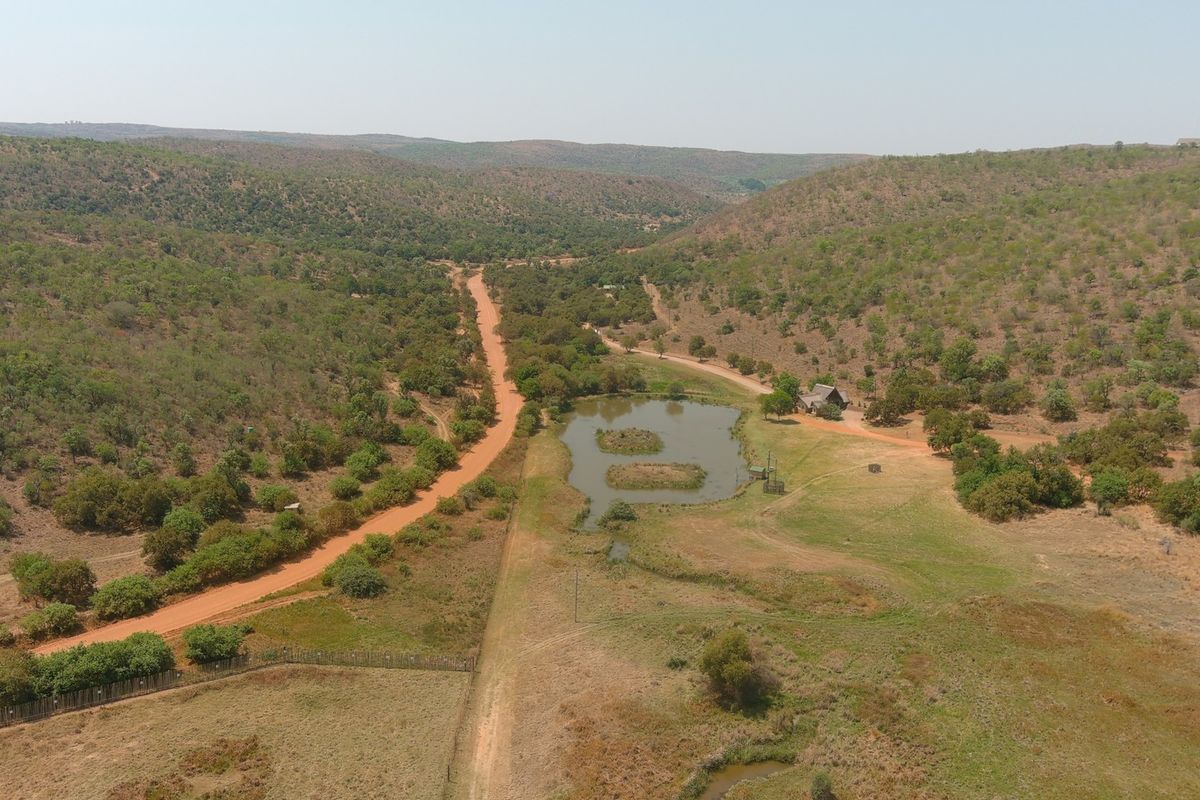 An aerial view of a rural property featuring a dirt access road, a natural dam, and expansive surrounding bushveld landscape.