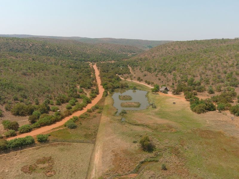 An aerial view of a rural property featuring a dirt access road, a natural dam, and expansive surrounding bushveld landscape.