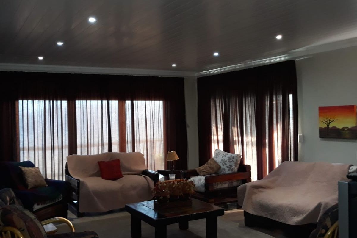 A spacious living room featuring a tongue and groove ceiling with recessed downlighting and large windows covered by dark curtains.