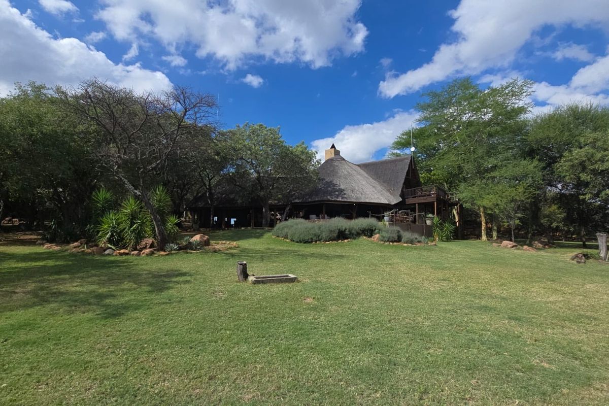 A view of the lodge exterior featuring a traditional thatched roof, expansive lawn, and mature trees in a natural bushveld setting.