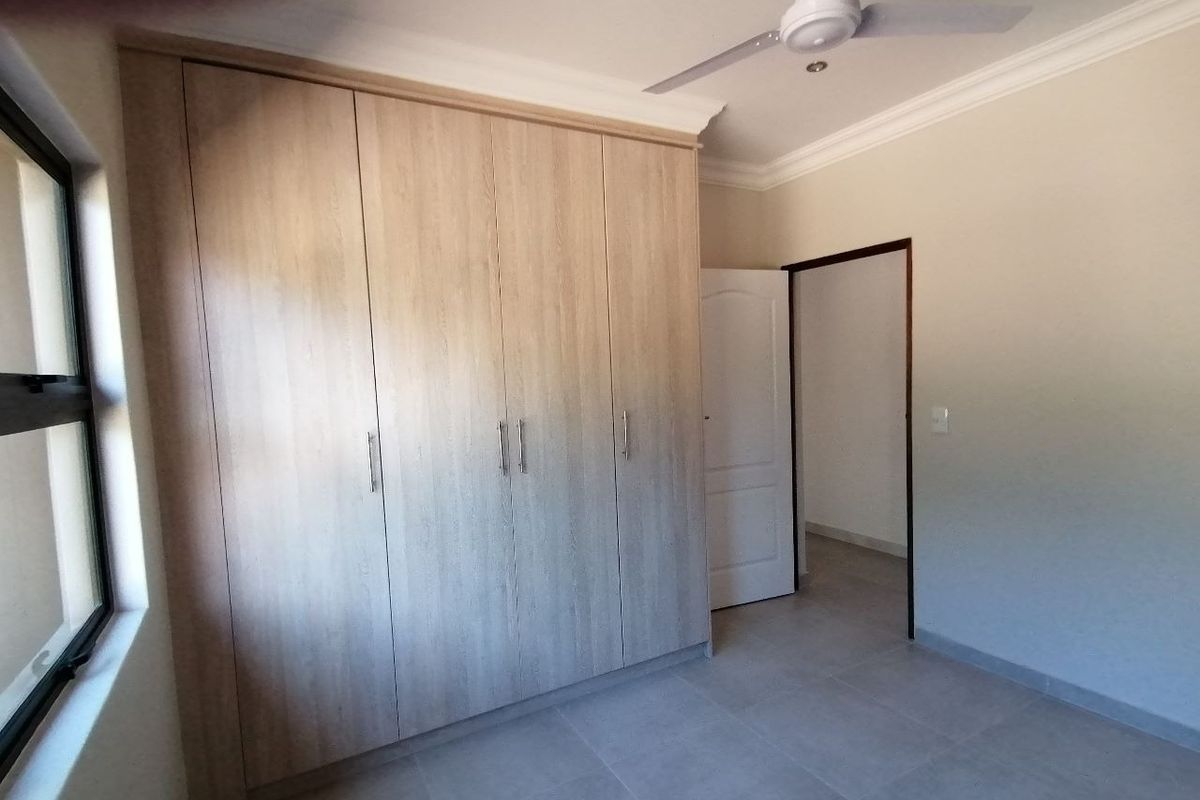 A clean, empty bedroom featuring modern light wood-finish built-in cupboards, tiled flooring, and a ceiling fan.