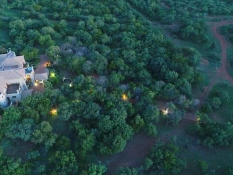 Aerial view of a lodge property nestled within a wooded landscape, featuring a tiled roof structure, swimming pool, and surrounding natural bushveld.