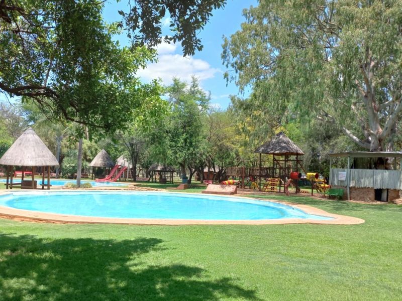 Outdoor lodge pool area featuring a swimming pool, multiple thatched gazebos, and a playground set within a spacious, well-maintained garden.