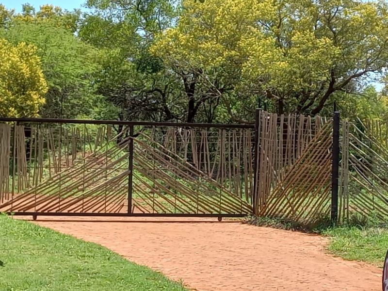 A rustic metal and bamboo entrance gate leading onto a paved driveway, surrounded by lush green trees and natural landscaping.