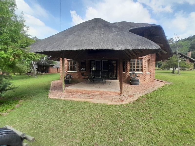 Rear view of a traditional farmhouse featuring a large thatched roof, exposed brick exterior, and a covered patio area set within a spacious garden.