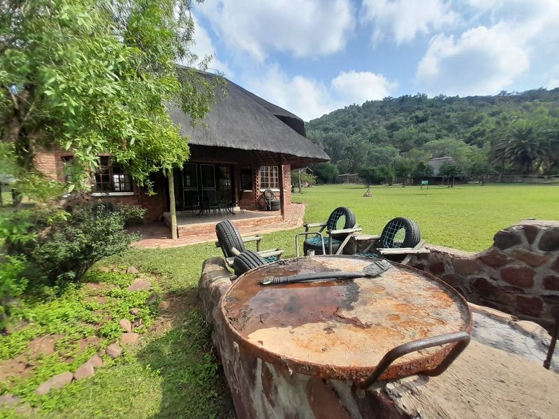 Rear view of a rustic farmhouse featuring a thatched roof, covered patio, and a stone-built outdoor braai area set within a spacious garden.