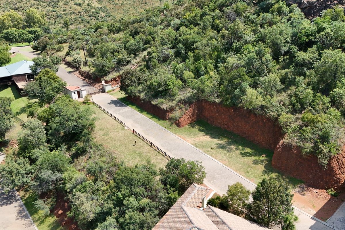 Aerial view of a residential vacant land plot featuring a paved driveway, security gatehouse, and natural hillside terrain with established vegetation.