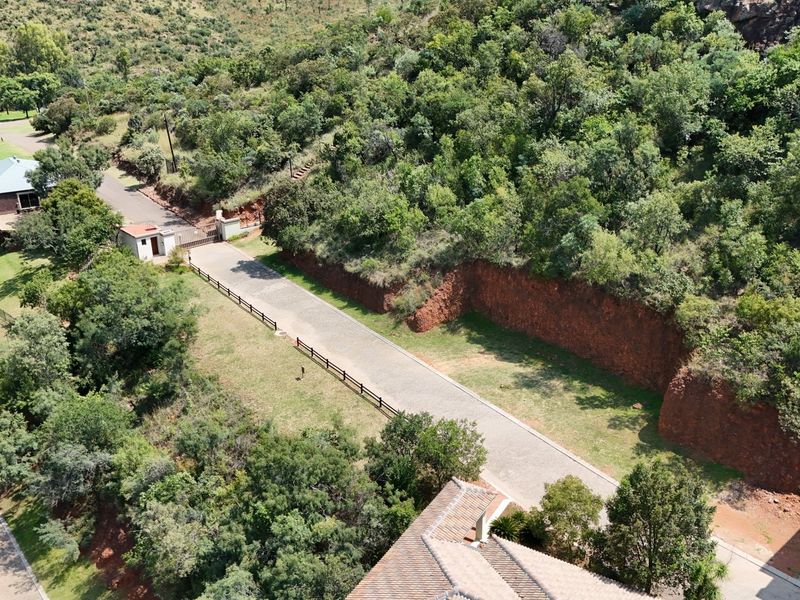 Aerial view of a residential vacant land plot featuring a paved driveway, security gatehouse, and natural hillside terrain with established vegetation.