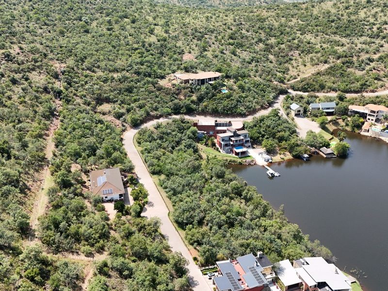 An aerial view of a residential estate featuring paved roads, native bushveld vegetation, and direct waterfront access to a dam or lake.
