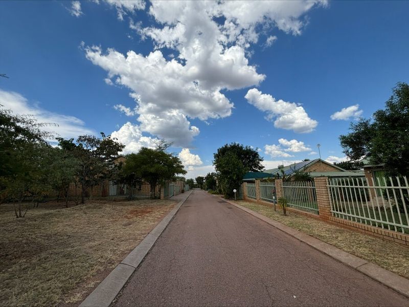 A view of a paved residential street lined with brick boundary walls, metal security fencing, and mature trees under a clear blue sky.