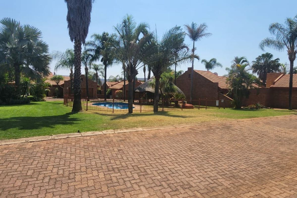 A wide view of the townhouse complex grounds featuring a swimming pool, landscaped gardens, paved driveway, and brick-built residential structures.