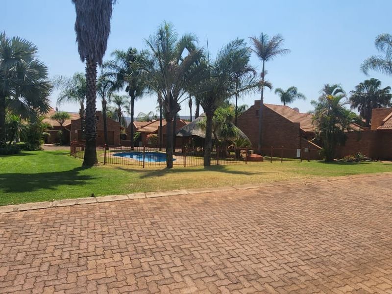 A wide view of the townhouse complex grounds featuring a swimming pool, landscaped gardens, paved driveway, and brick-built residential structures.