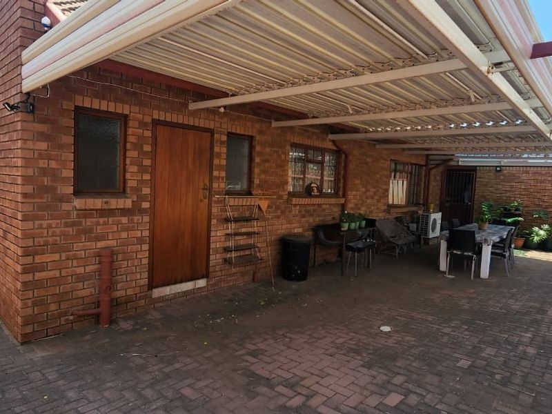 A paved rear patio area featuring a large metal-framed covered pergola, brick exterior walls, and access to the main dwelling.