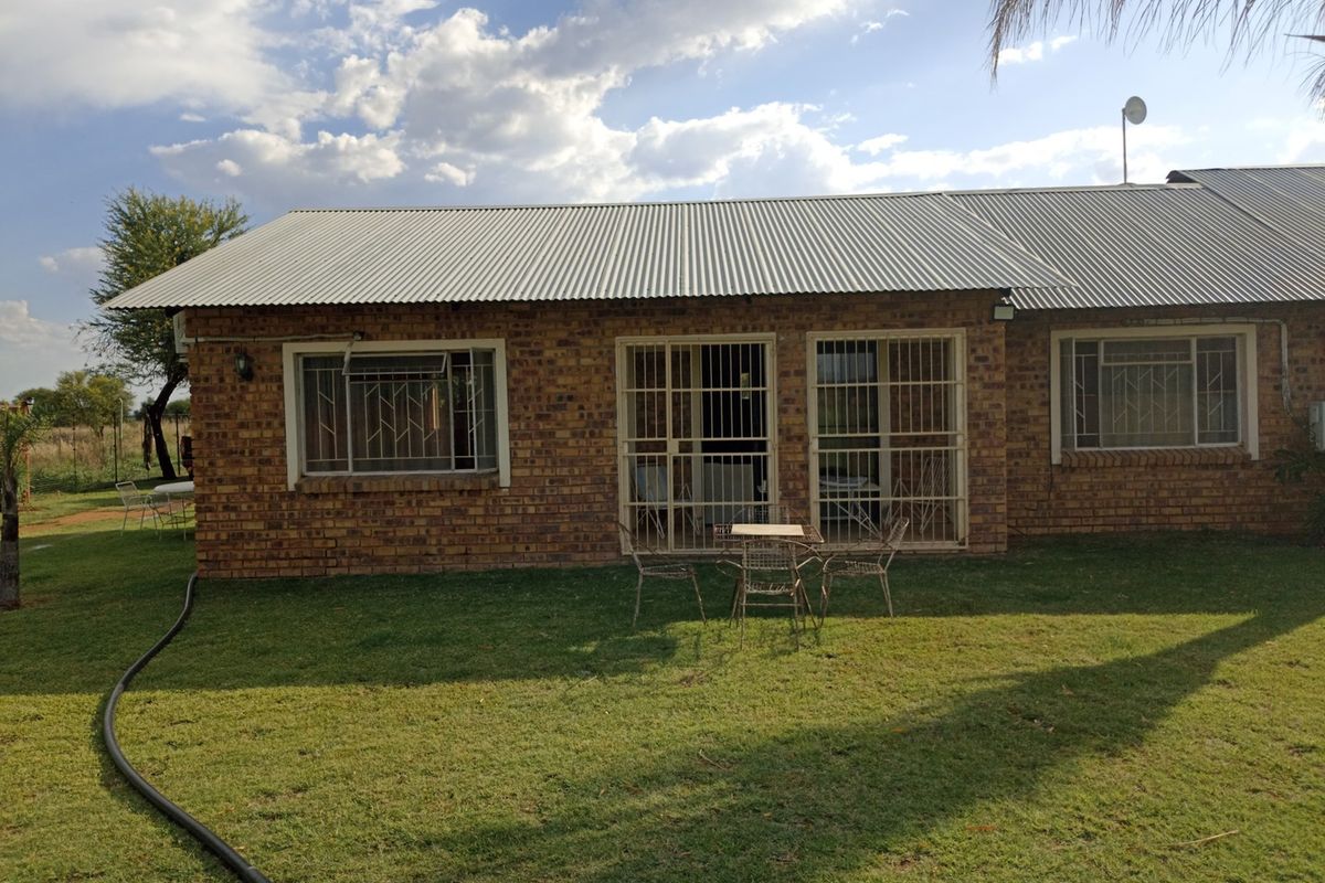 Rear view of a brick-built small holding dwelling featuring corrugated metal roofing and security-fitted windows and doors overlooking a garden area.