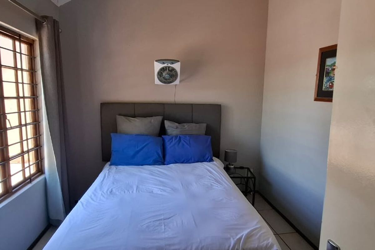 A compact bedroom featuring tiled flooring, a large window with security bars, and a wall-mounted fan.