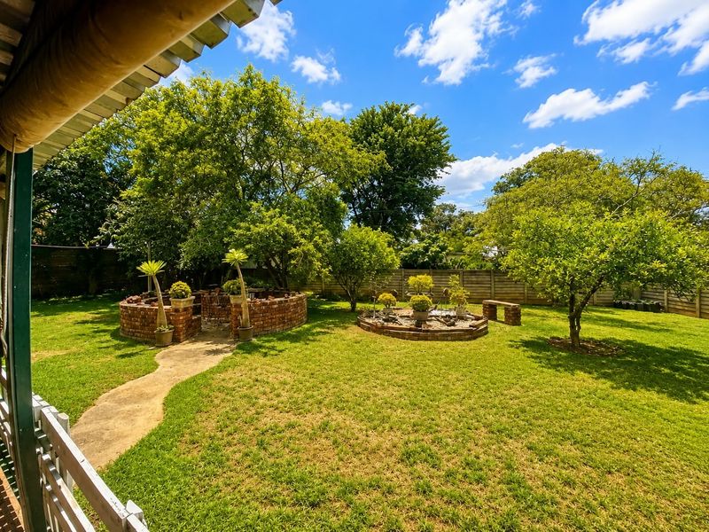 A well-maintained garden featuring a lush lawn, mature trees, a circular brick retaining wall, and a concrete pathway, enclosed by a precast boundary wall.