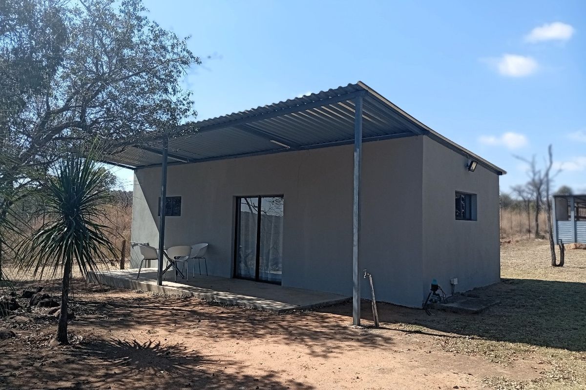 A simple farm-style dwelling featuring a covered patio, corrugated metal roof, and sliding glass door access, set in a natural rural landscape.