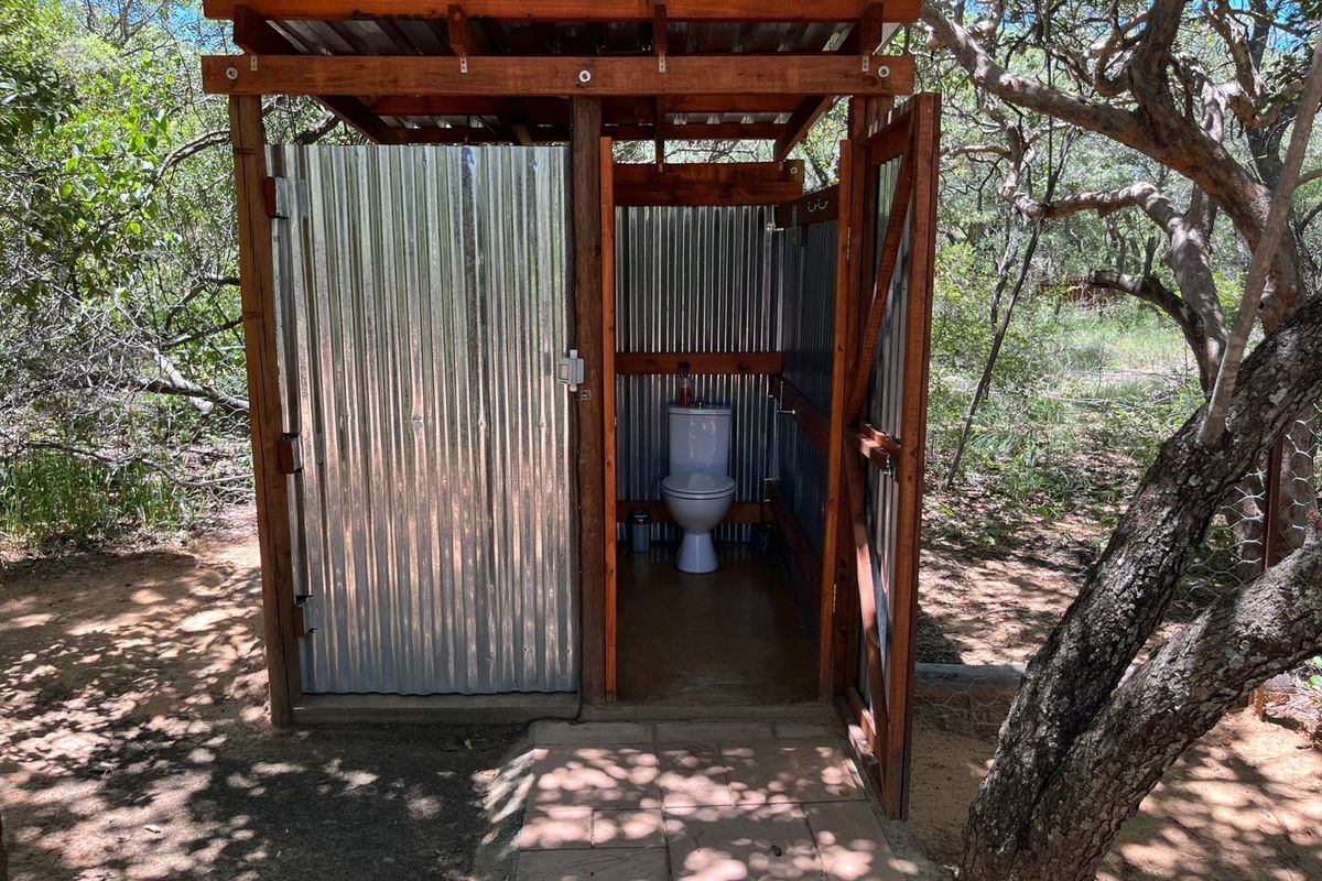 An outdoor rustic bathroom structure featuring corrugated metal walls, a timber frame, and a standard ceramic toilet, set within a natural wooded environment.