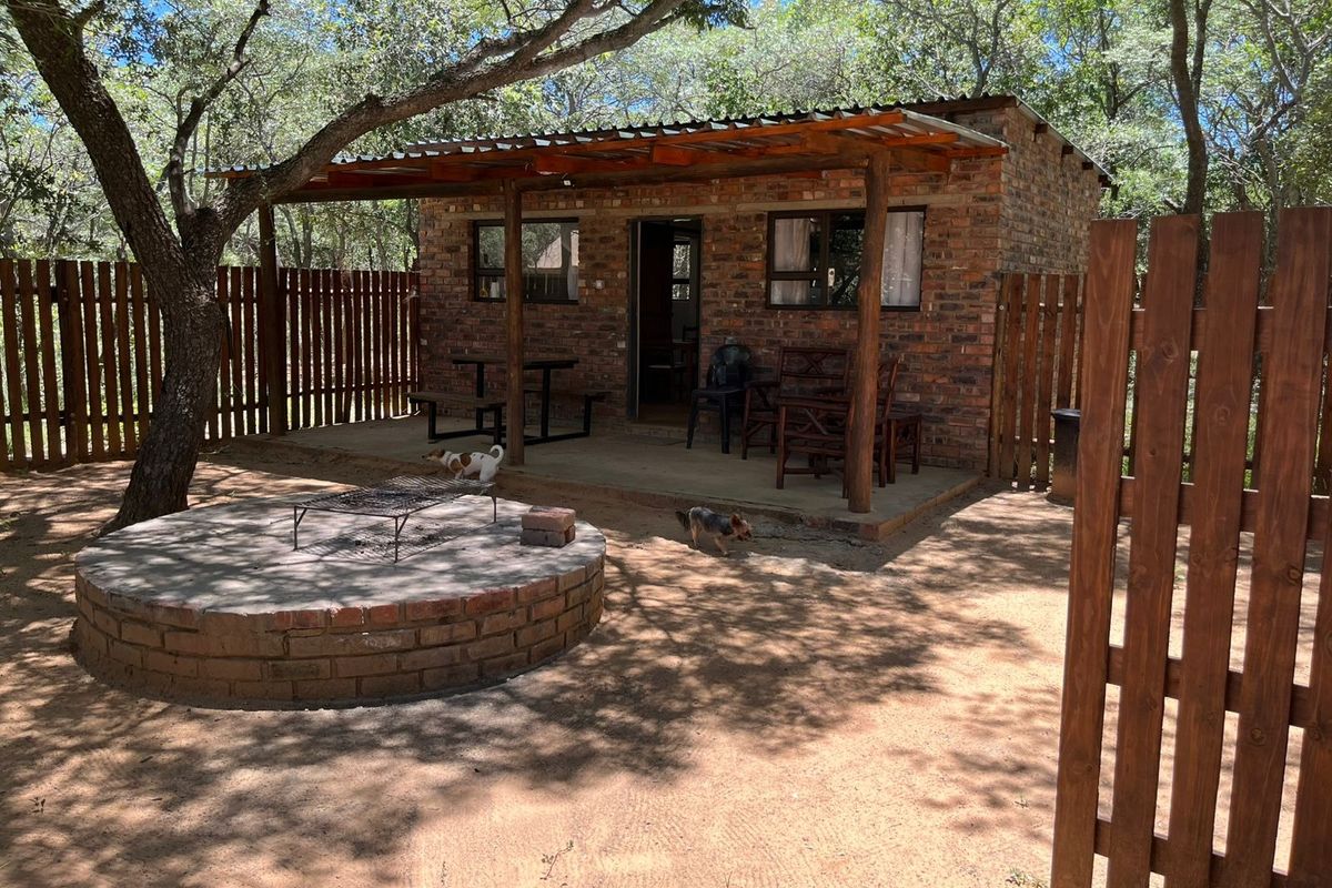A rustic brick cottage featuring a covered patio area and a built-in circular brick fire pit, set within a natural wooded farm environment.