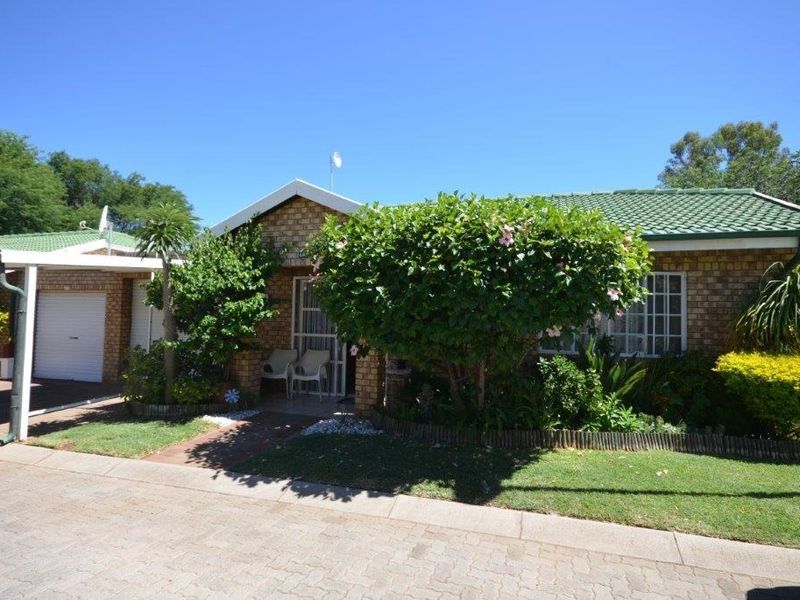 Front elevation of a face brick retirement unit featuring a green tiled roof, attached carport, and established garden with paved access.