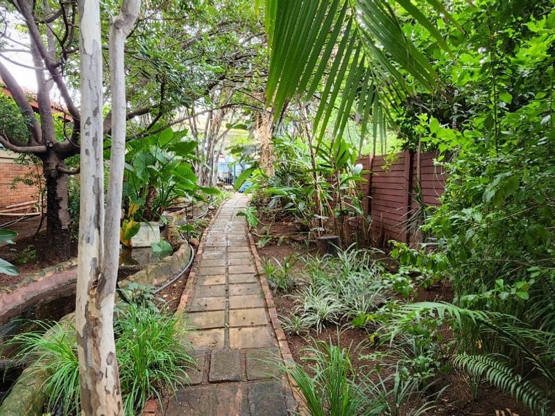 A lush, established garden path featuring a brick walkway, mature trees, and a built-in water feature alongside the property's exterior brick wall.