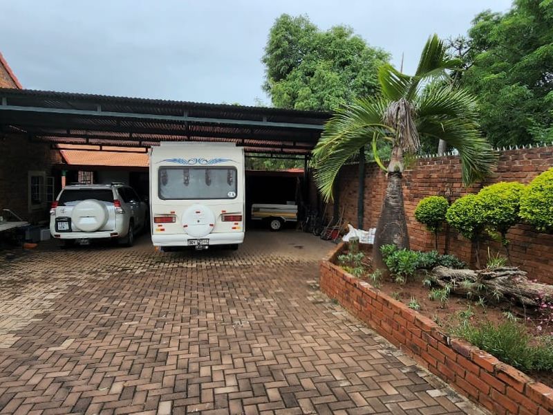 Front exterior view featuring a brick house with a large covered carport, paved driveway, and a landscaped garden bed along the boundary wall.