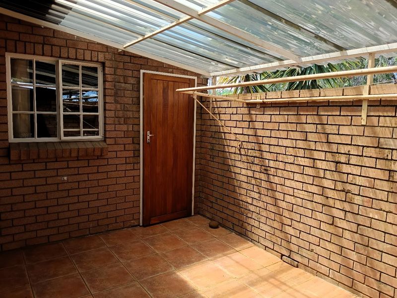 A covered outdoor patio area featuring exposed brick walls, tiled flooring, and a translucent roof, providing a functional space for laundry or relaxation.