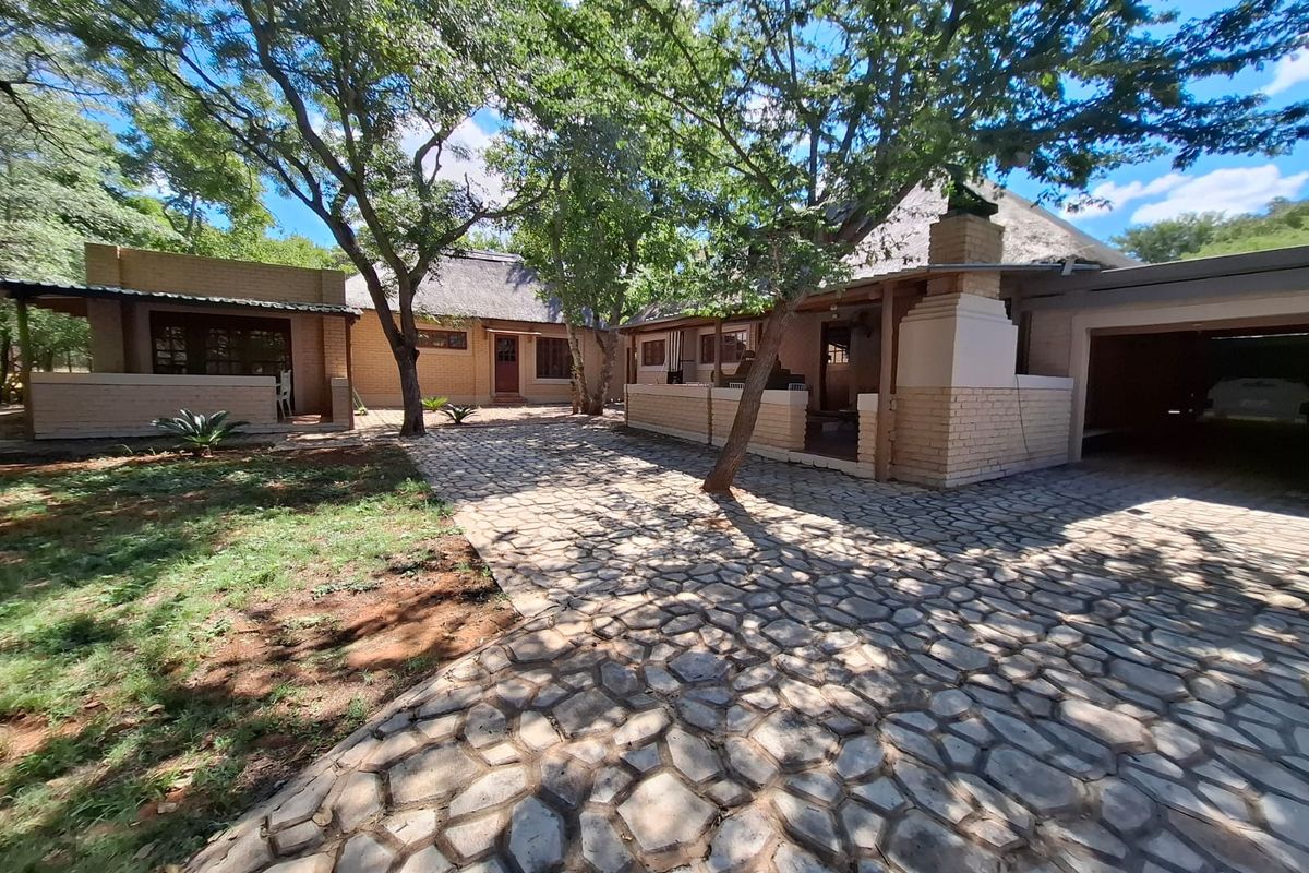 A spacious rear courtyard featuring stone paving, mature trees, a thatched roof dwelling, and a built-in outdoor fireplace.