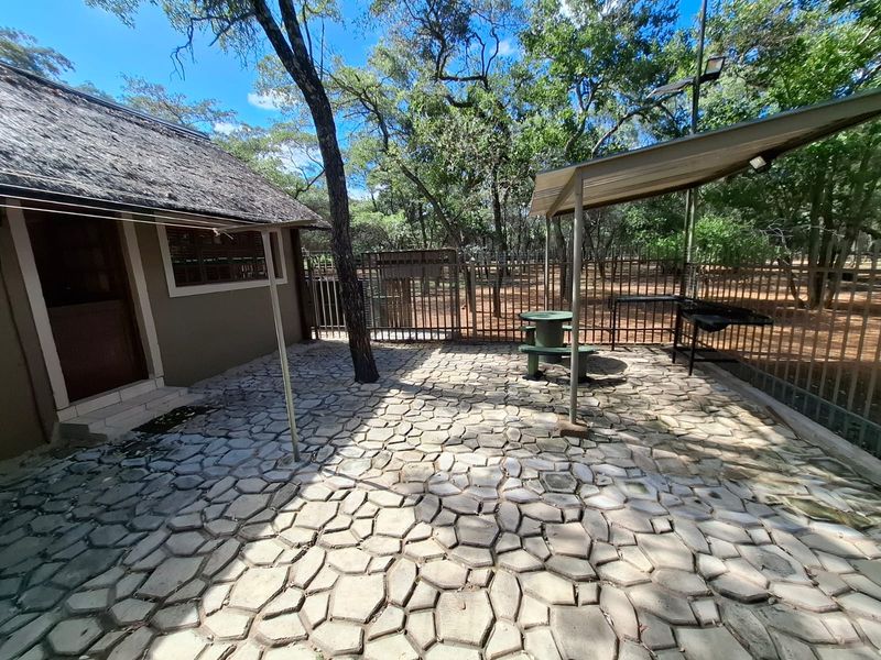 A covered outdoor patio area featuring paved flooring and metal fencing, set within a wooded garden environment.
