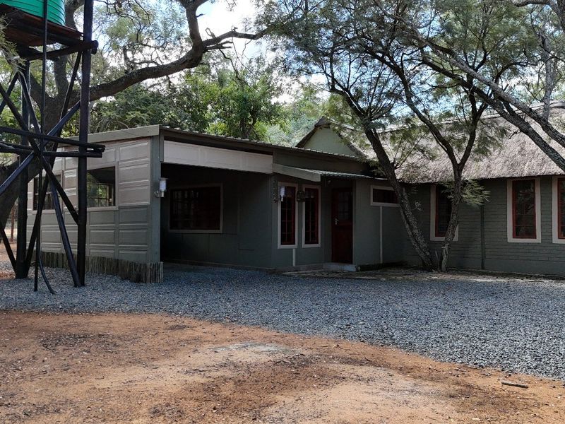 Front view of a small holding residence featuring a thatched roof, attached carport, and a prominent elevated water tank structure on a gravel driveway.