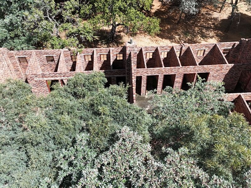 Aerial view of a brick structure with an exposed roof framework, surrounded by mature trees on a small holding property.