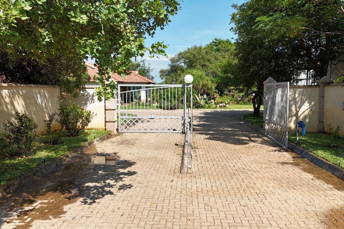 A gated residential entrance featuring a paved driveway, perimeter walls, and metal security gates leading into the property grounds.