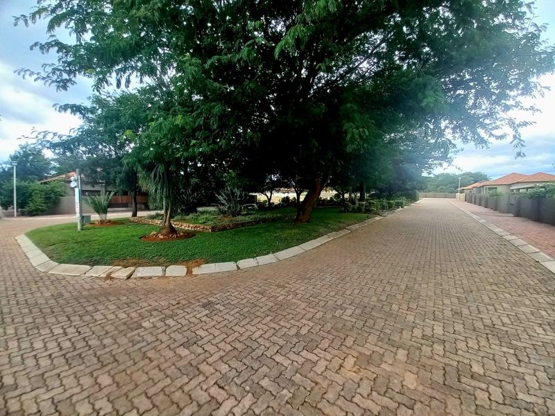 A view of the paved residential street and driveway, featuring mature trees and well-maintained landscaped garden areas within the estate.