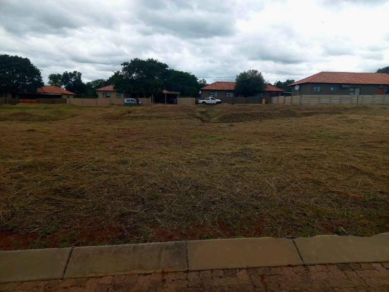 A vacant residential plot featuring a cleared grass surface, bordered by a paved curb and surrounding boundary walls in a developed neighborhood.
