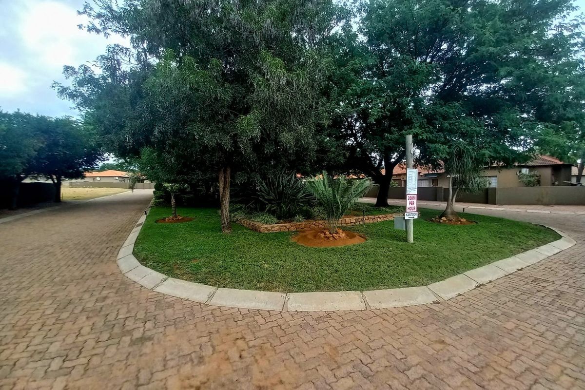 A well-maintained residential street view featuring a paved driveway, mature trees, and landscaped garden areas within a residential estate.