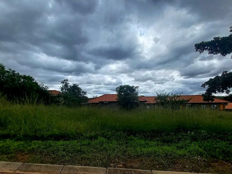 A view of a vacant residential plot featuring overgrown grass with surrounding residential buildings in the background under a cloudy sky.