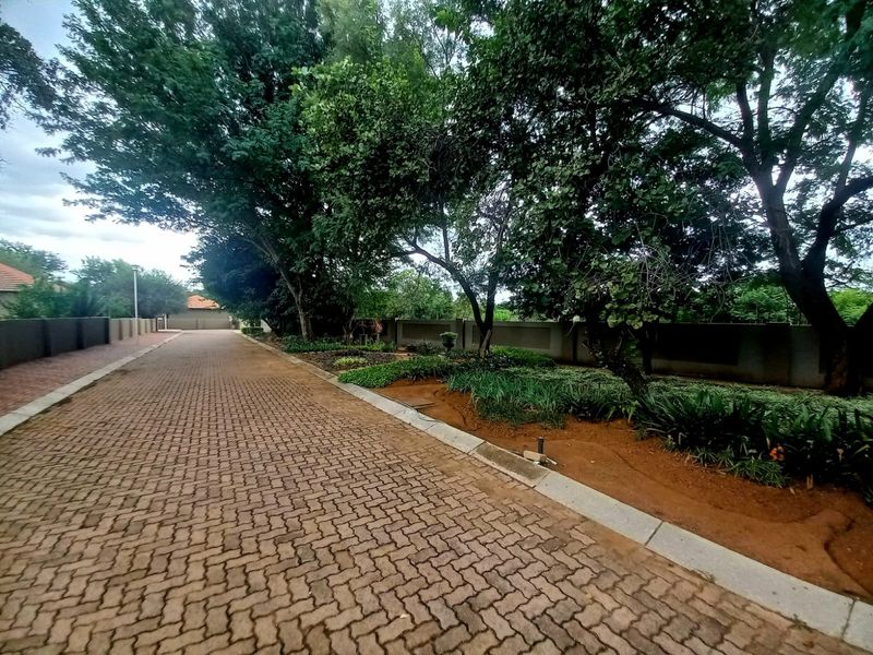 A well-maintained paved driveway leading through a landscaped garden area with mature trees and a secure perimeter wall.