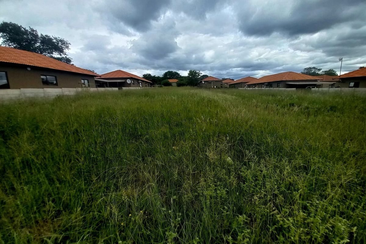 A vacant residential plot featuring overgrown grass, enclosed by perimeter walls and situated within a developed residential estate.