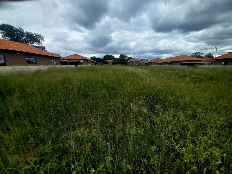 A vacant residential plot featuring overgrown grass, enclosed by perimeter walls and situated within a developed residential estate.