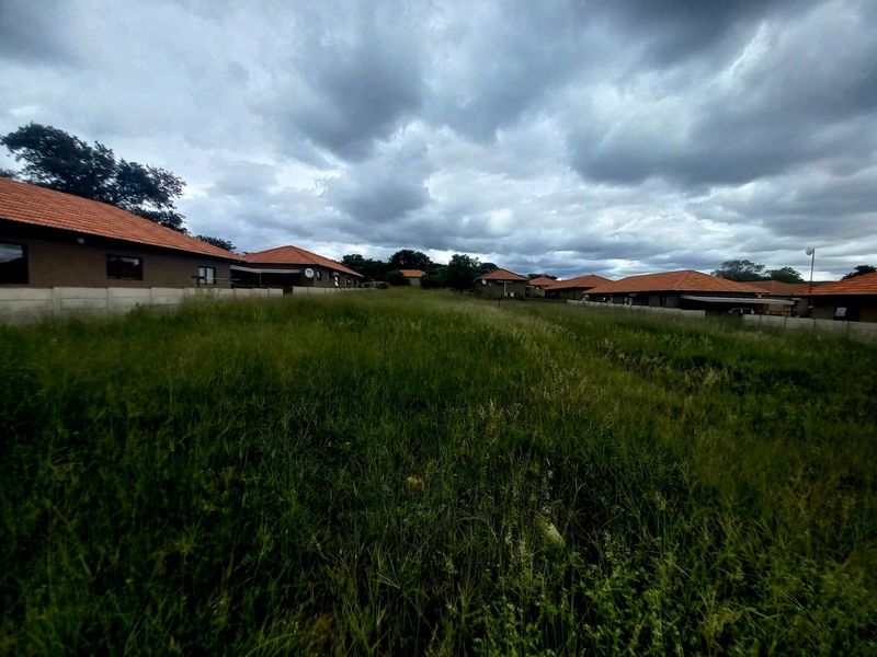 A view of a vacant residential plot featuring overgrown grass, enclosed by perimeter boundary walls with neighboring residential properties visible in the background.