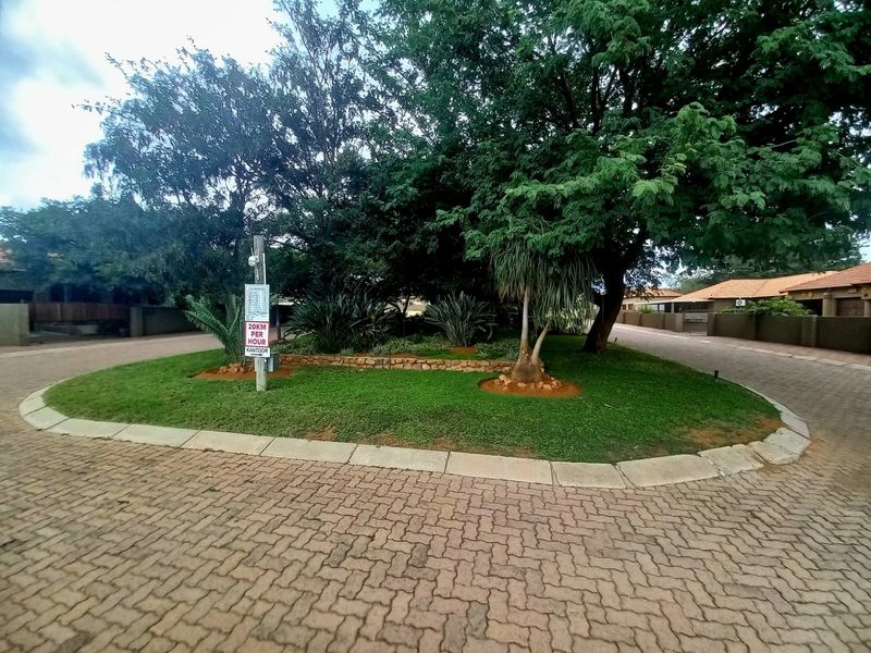 A well-maintained residential street view featuring a paved driveway, landscaped garden island with mature trees, and a stone retaining wall.