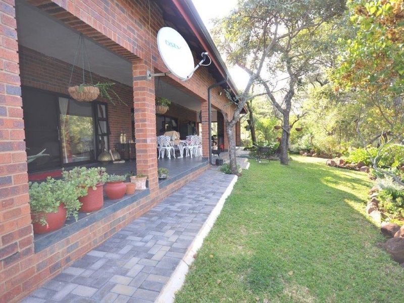 A spacious covered brick patio featuring tiled flooring and sliding door access, overlooking a well-maintained garden with mature trees and paved pathways.