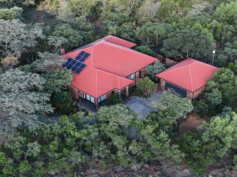 Aerial view of a brick house with a red tiled roof, solar panels, and a paved driveway, nestled within a lush, wooded landscape.