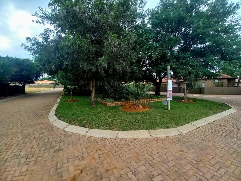 A well-maintained residential street view featuring a paved driveway, mature trees, and landscaped garden areas within a residential estate.