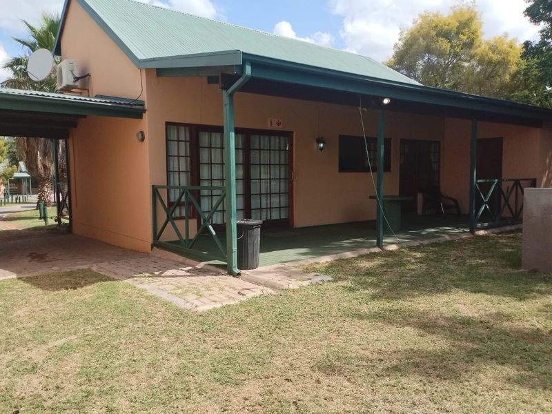 Rear view of the apartment featuring a covered patio area, metal roofing, and paved access, set within a landscaped garden environment.