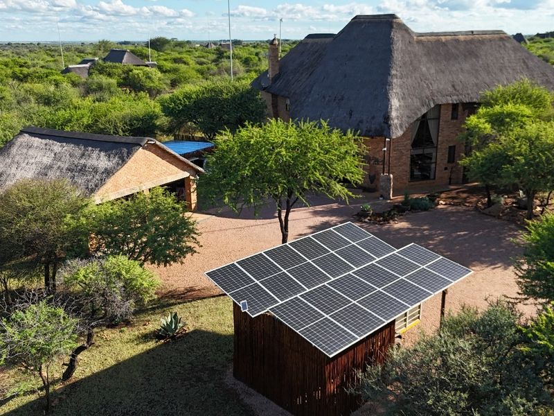 Aerial view of a guest house property featuring traditional thatch roof buildings, a gravel driveway, and a large integrated solar panel array.