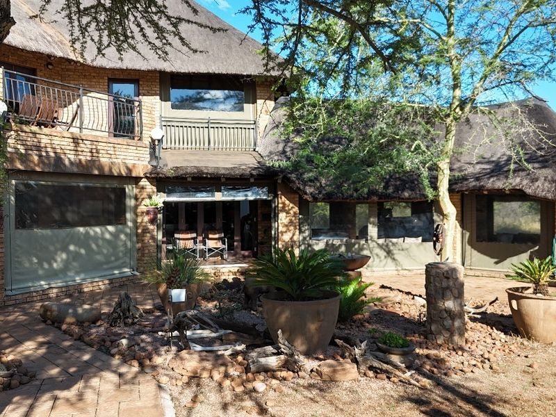Rear view of a guest house featuring a traditional thatch roof, brick exterior, upper-level balcony, and a landscaped patio area.
