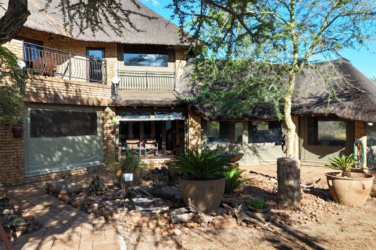 Rear view of a guest house featuring a traditional thatch roof, brick exterior, upper-level balcony, and a landscaped patio area.