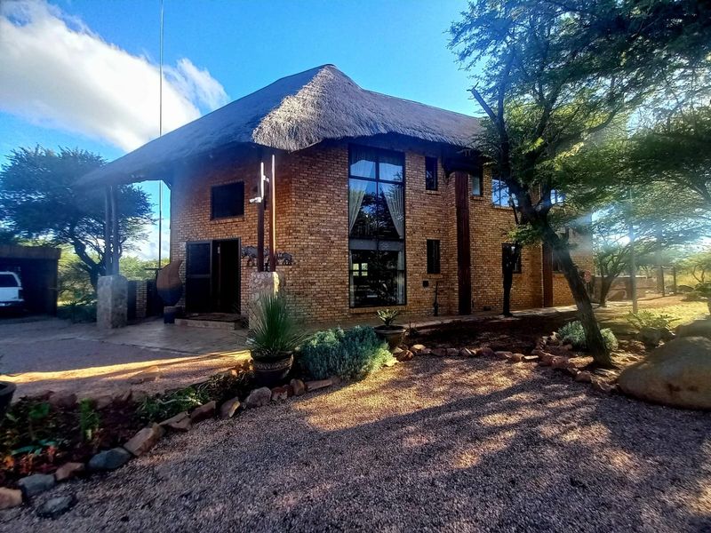 A charming double-story guest house featuring a traditional thatch roof, face brick exterior, and large windows set within a natural, landscaped garden environment.