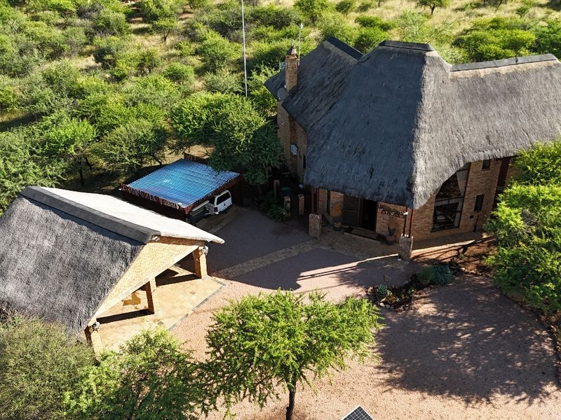 Aerial view of a guest house property featuring traditional thatched roofs, brick construction, a paved driveway, and a detached carport set in a natural landscape.